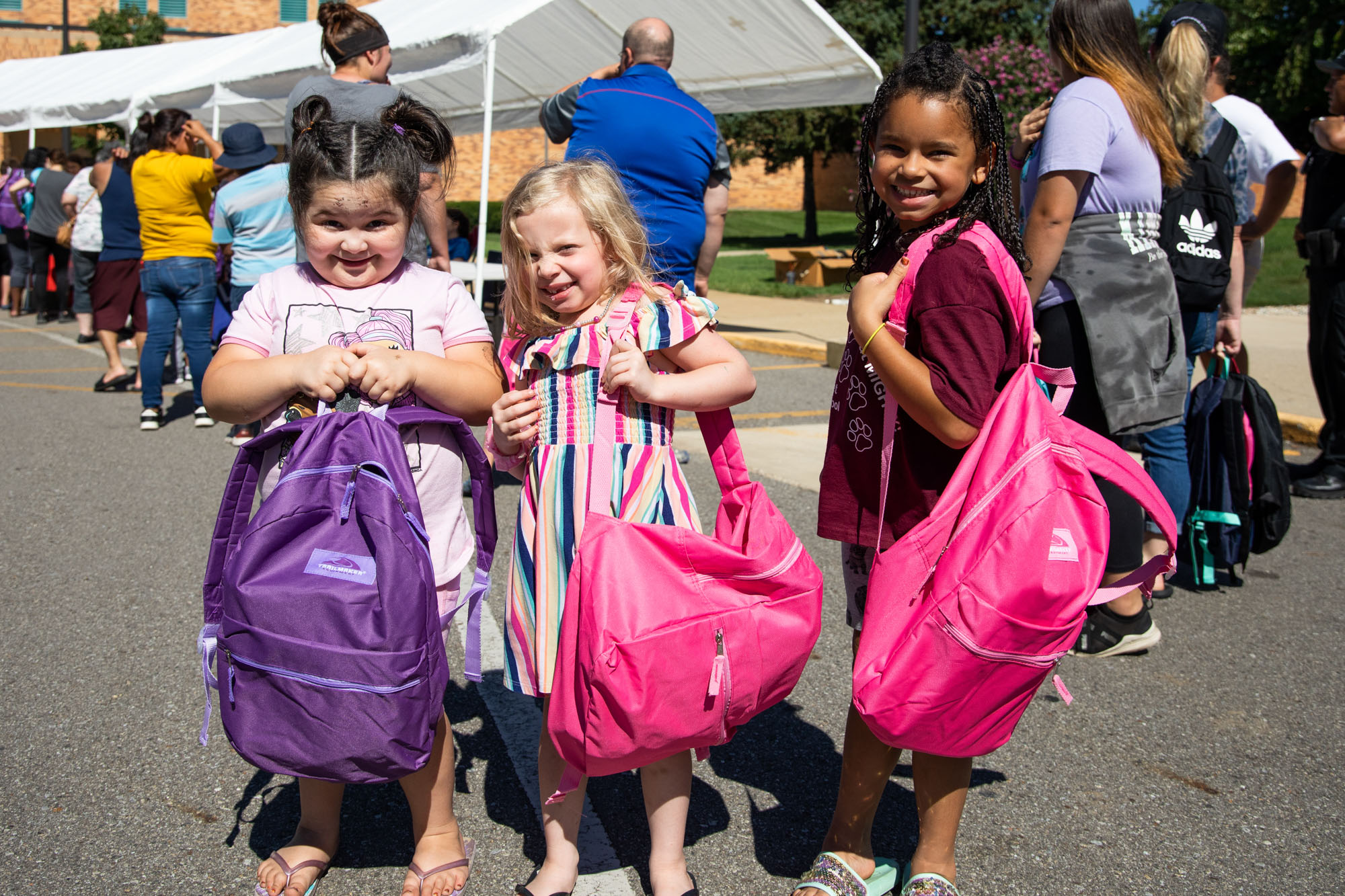 Three young girls holding up their pink bookbags. The girl on the left is holding a purple backpack.