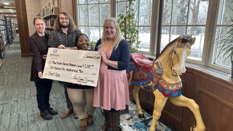 A group of four people standing near a window and decommissioned carousel horse while holding a large check inside the Maud Preston Palenske Luibrary.